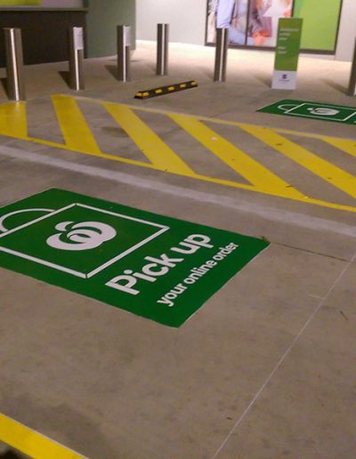 Two Woolworths carparks outside a shopping centre for customers to park when collecting online orders. Two carparks have a walkway painted between them and a sign in the middle of each with the Woolworths logo and Pick up your online order.